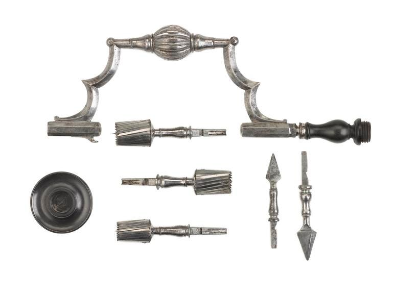 A modern photograph of a set of metal surgical tools. There is a drill-like device at the top of the image, three circular drill bits of differing sizes, two pointed attachments and a round dark-coloured handle.