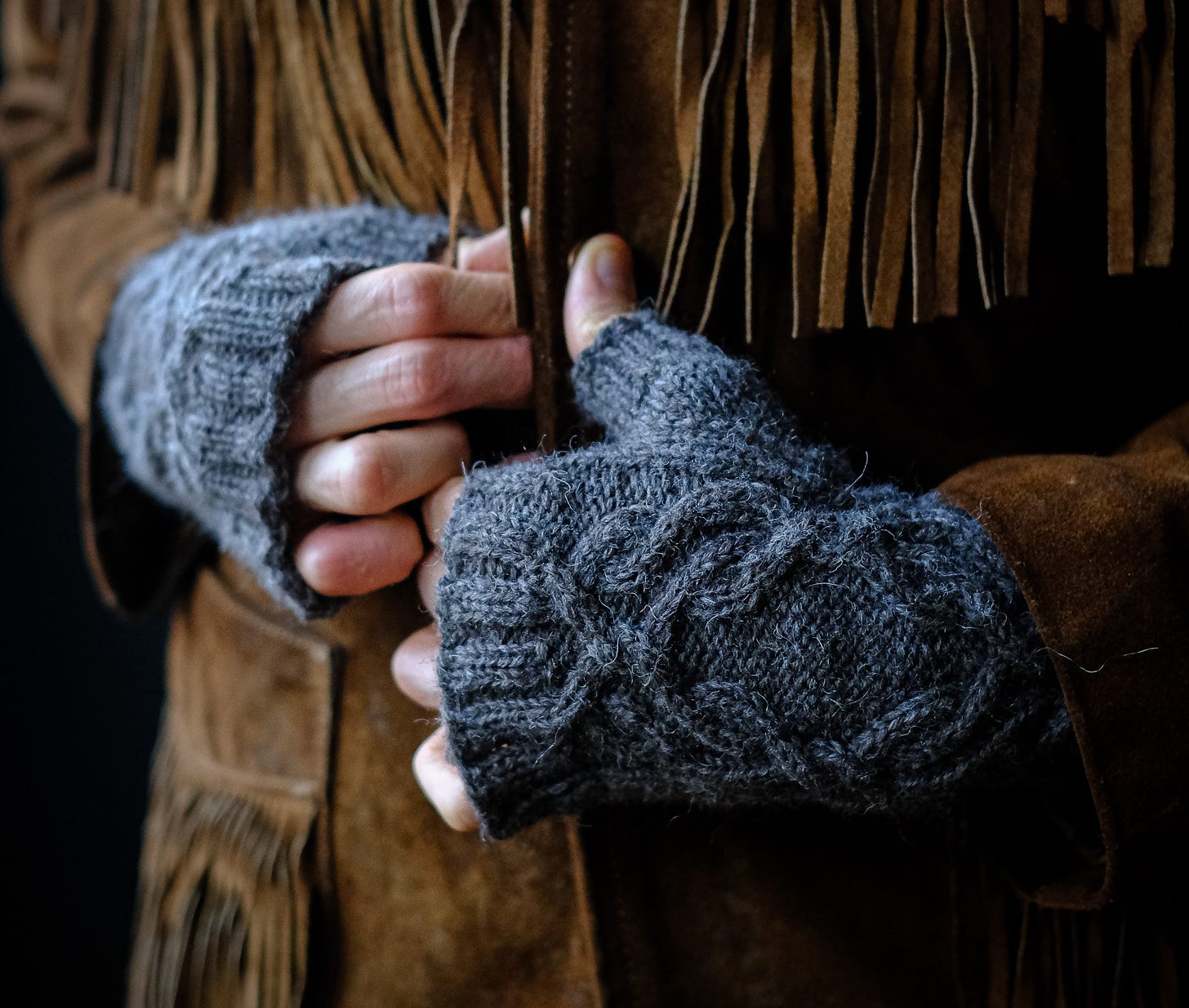 Two hands, wearing a pair of handknitted, cabled fingerless mittens close the final clasp of a suede jacket. Fringe hanging from the breast of the jacket tickling and caressing the fingers of our subject as the cool, evening light pours in from the left. The wool is sticky, textured and rough. Fisherman wool. Woodworker. Wool made for stability and security. In the distance, the smell of pine lingers in the damp air — carried by the fog rolling in off the mountains. Foxes feet silently running through the undergrowth. The soft cry of an elk echoing. 