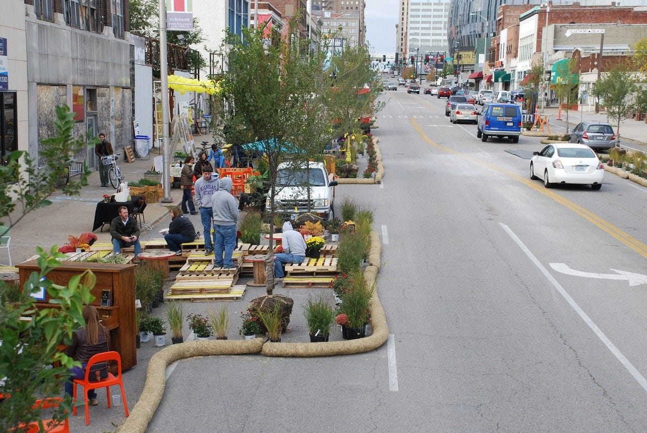 In a city street with four wide lanes of traffic, Better Block sets up "parklets" in what are now parking spaces. Hugging the sidewalk are pallets for sitting, trees and shrubs in their pre-planting pots, chairs, tables, and a piano, all surrounded by sand bag "guard rails."