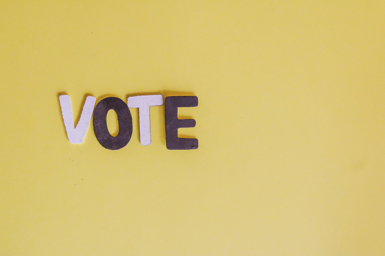 La parola "Vote" con lettere rosa e viola appoggiate su uno sfondo giallo ocra.