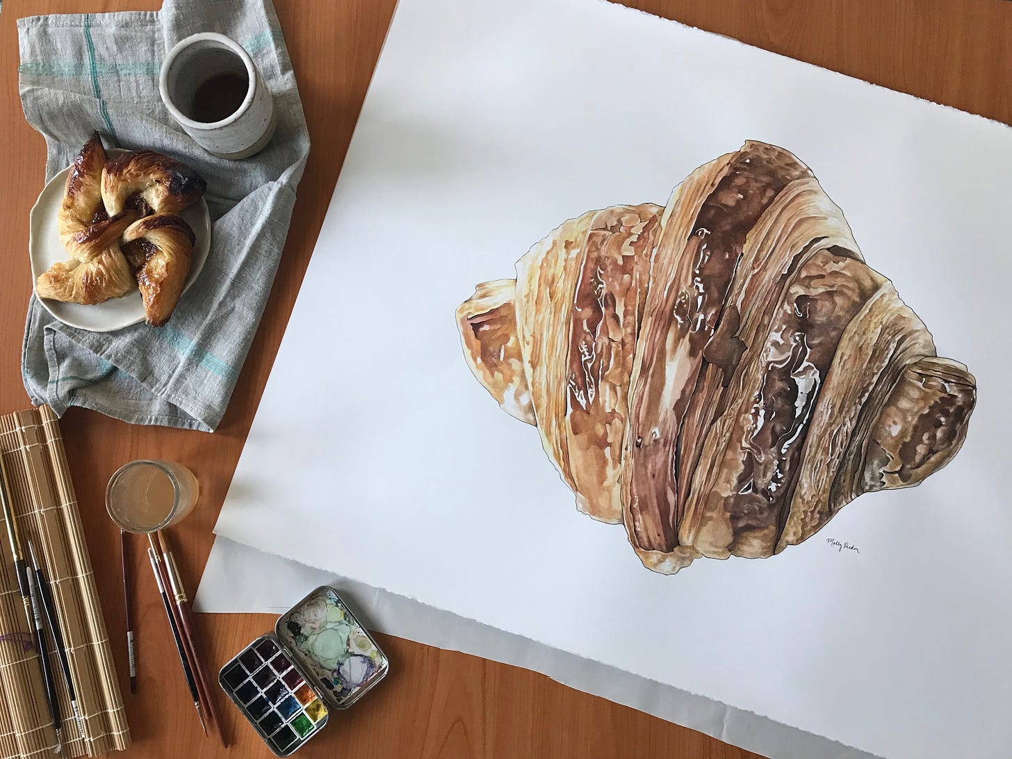 Birds eye view of the painting on a desk surface with a croissant pastry to the left of it, a cup of coffee, and painting supplies