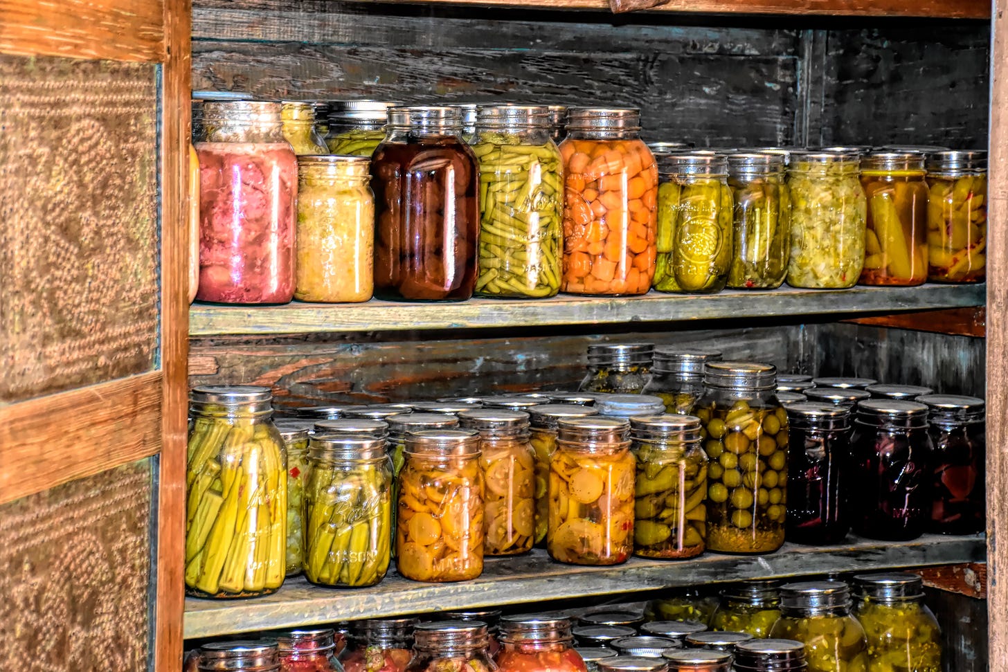 Two wooden shelves are stacked with dozens of glass jars filled with pickles and preserves