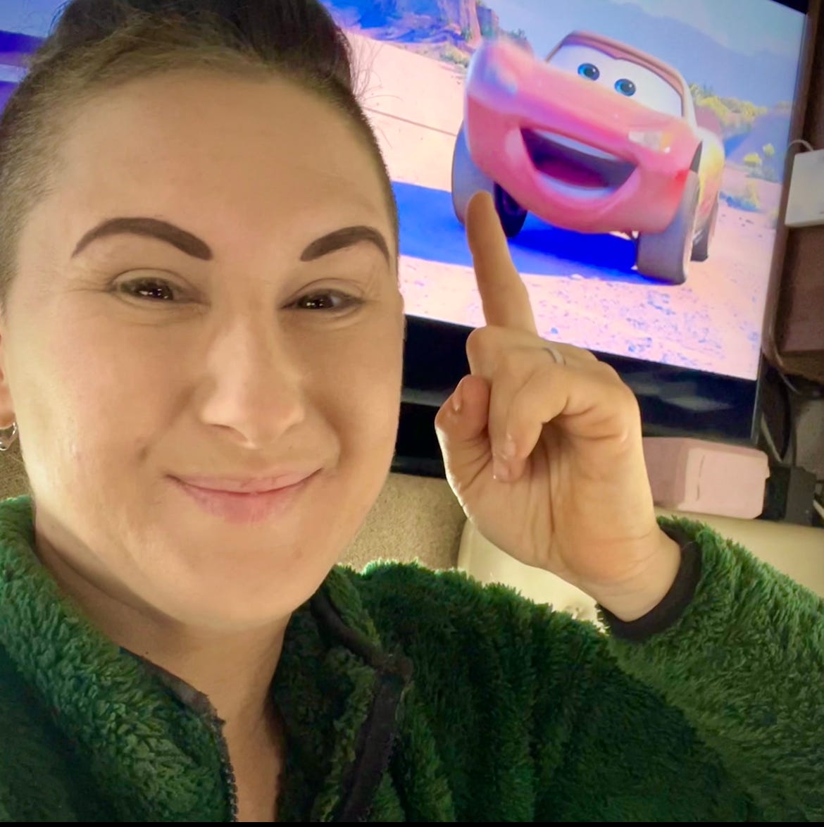 Photo of Lyric, a fair skinned multiracial human in their mid 30s, with short hair and shaved sides, in their RV, in front of a TV that has Disney's Cars and Lightning McQueen smiling on the screen behind them.