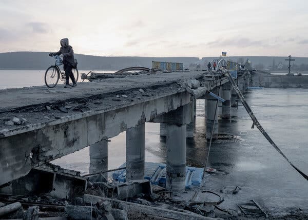 A warmly dressed woman with a bicycle crosses a badly damaged bridge.