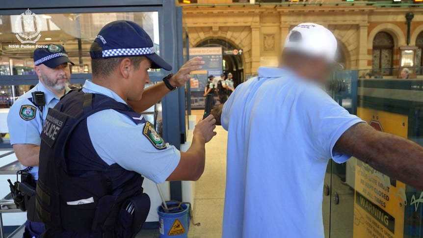 A male police officer searches a man inside a train station using an electronic scanner.