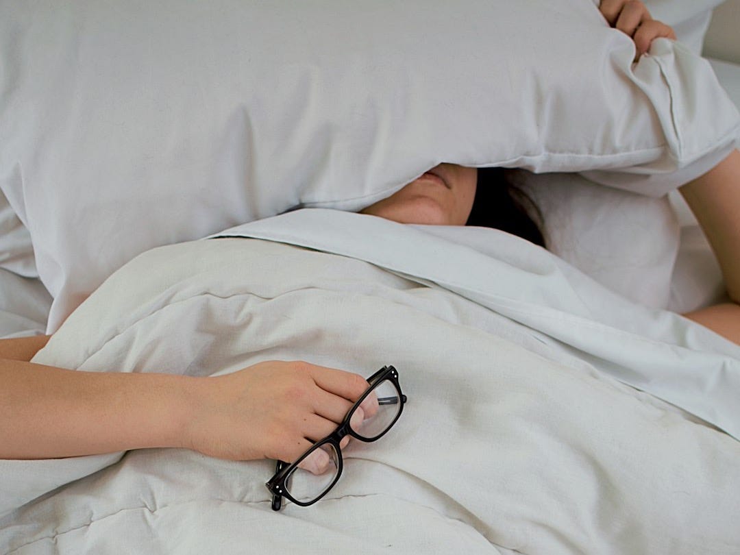 person lying on bed while covering face with pillow and holding eyeglasses