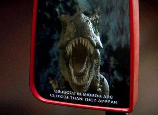 Image: A t-rex growls into a side view mirror that says "Objects in mirror are closer than they appear"