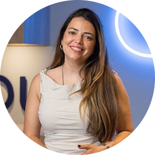 A smiling woman with long brown hair wearing a white sleeveless top stands in front of a blue and white background with part of a company logo visible.