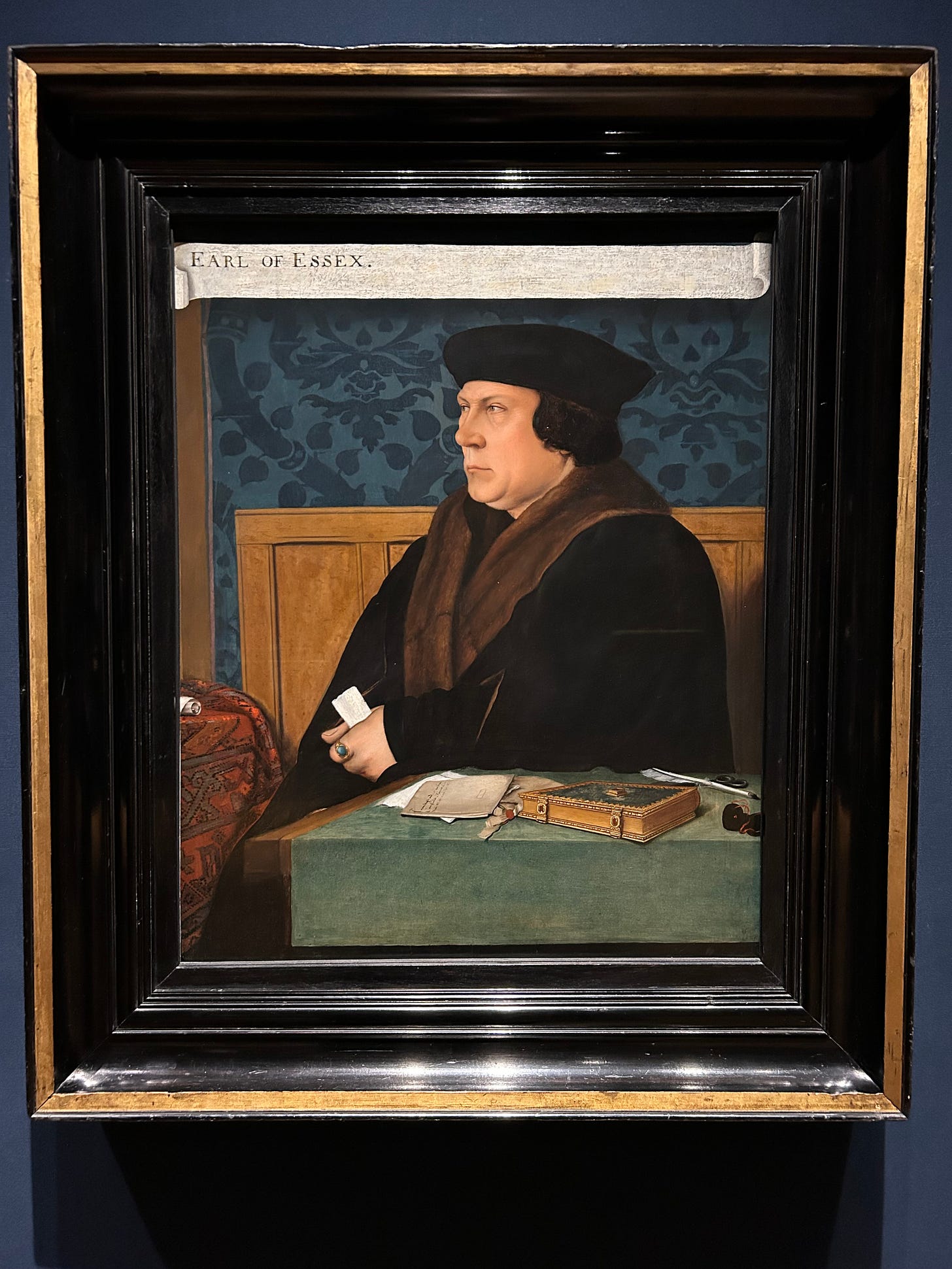 Thomas Cromwell after Hans Holbein. This middle-aged man sits behind a desk that contains correspondence, a green book, a quill pen, ink, and a black drawstring purse. He clutches a document in his fist. 