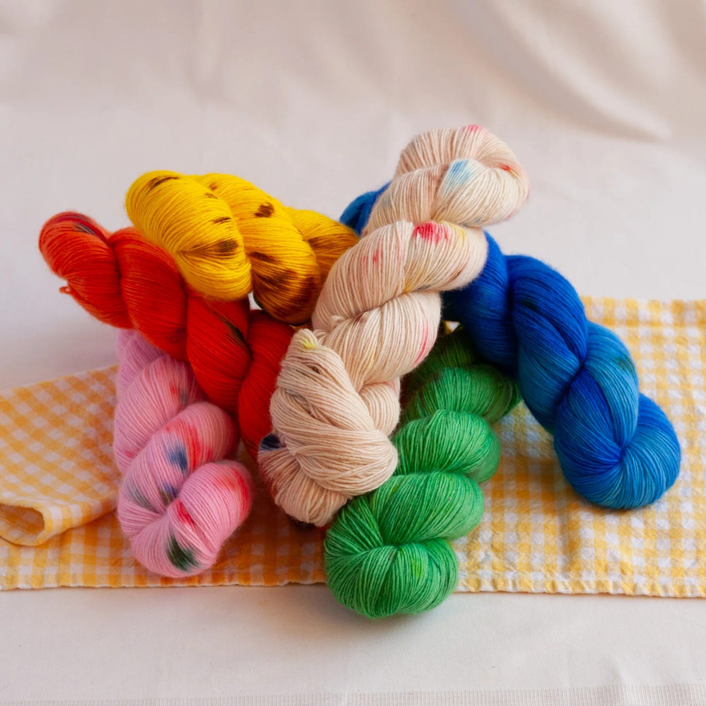 A pile of hand dyed yarn thrown casually on a gingham cloth. The yarns are bright red, yellow, blue, pink, and white with colourful speckles. 