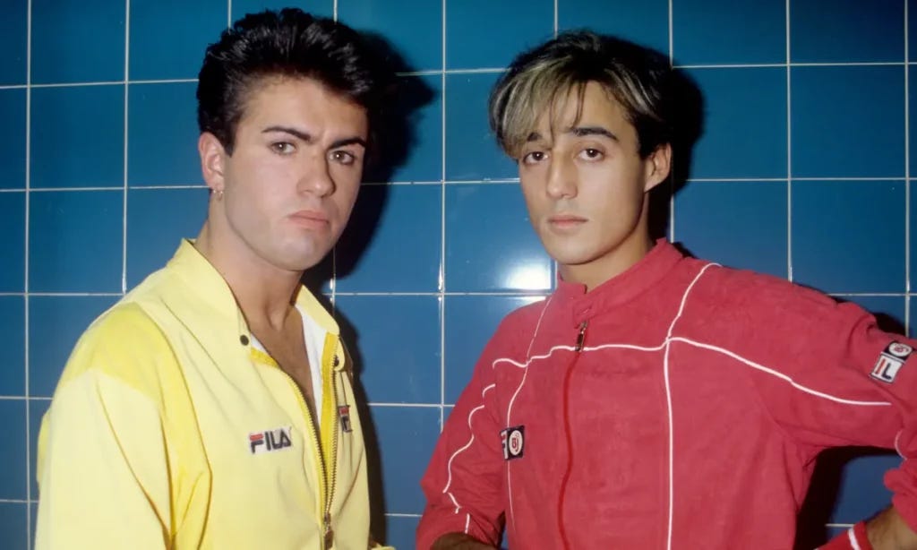 Two handsome, young men stand in front of a blue square tile wall wearing 80s Fila sports jackets in primary yellow and red.