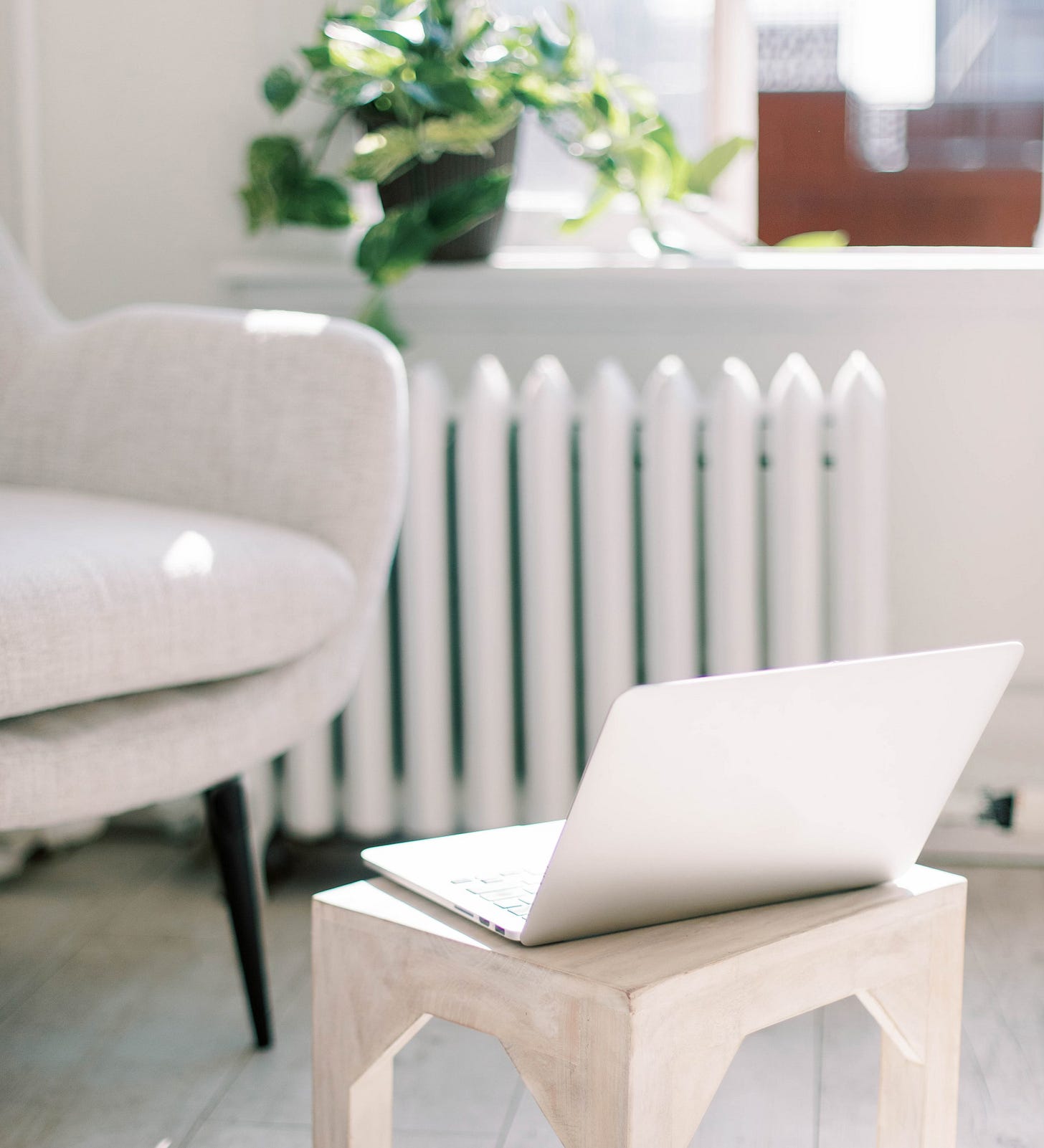 A minimal, sunlit workspace with a white laptop on a small wooden stool beside a neutral armchair. Natural light filters through the window behind a green plant.