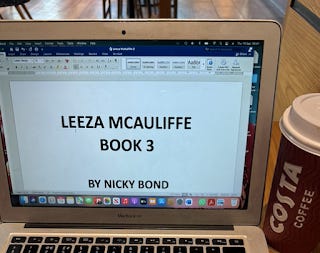 An open laptop on a coffee shop table. A tall take-away cup is next to the laptop. It's a maroon colour and has COSTA written down the side in white lettering. On the laptop screen, there's an open word doc. It has the words 'Leeza McAuliffe Book 3 by Nicky Bond' written in large black font.