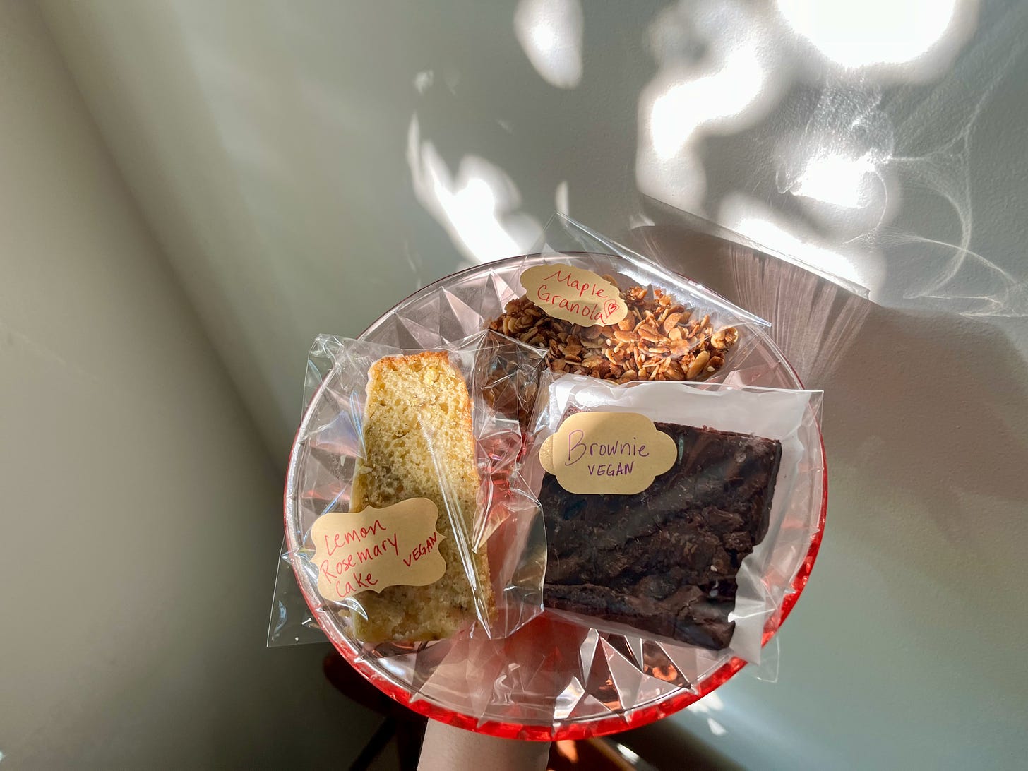 I hold three treats wrapped in cellophane bags on a cake plate: Lemon Rosemary Cake, Maple Granola, and a Brownie. The light is pretty.