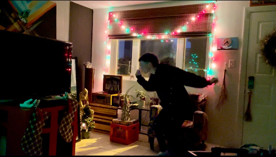 A photograph of Lyss dancing in their living room, in front of a window with Christmas lights. They are wearing a black track suit, and they are surrounded by various holiday decorations.
