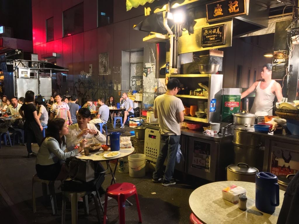 An image of a street food stall, with a cook, a server standing at his station and various customers who are seated and eating dishes on plastic chairs and formica tables in the middle of a pedestrianised street