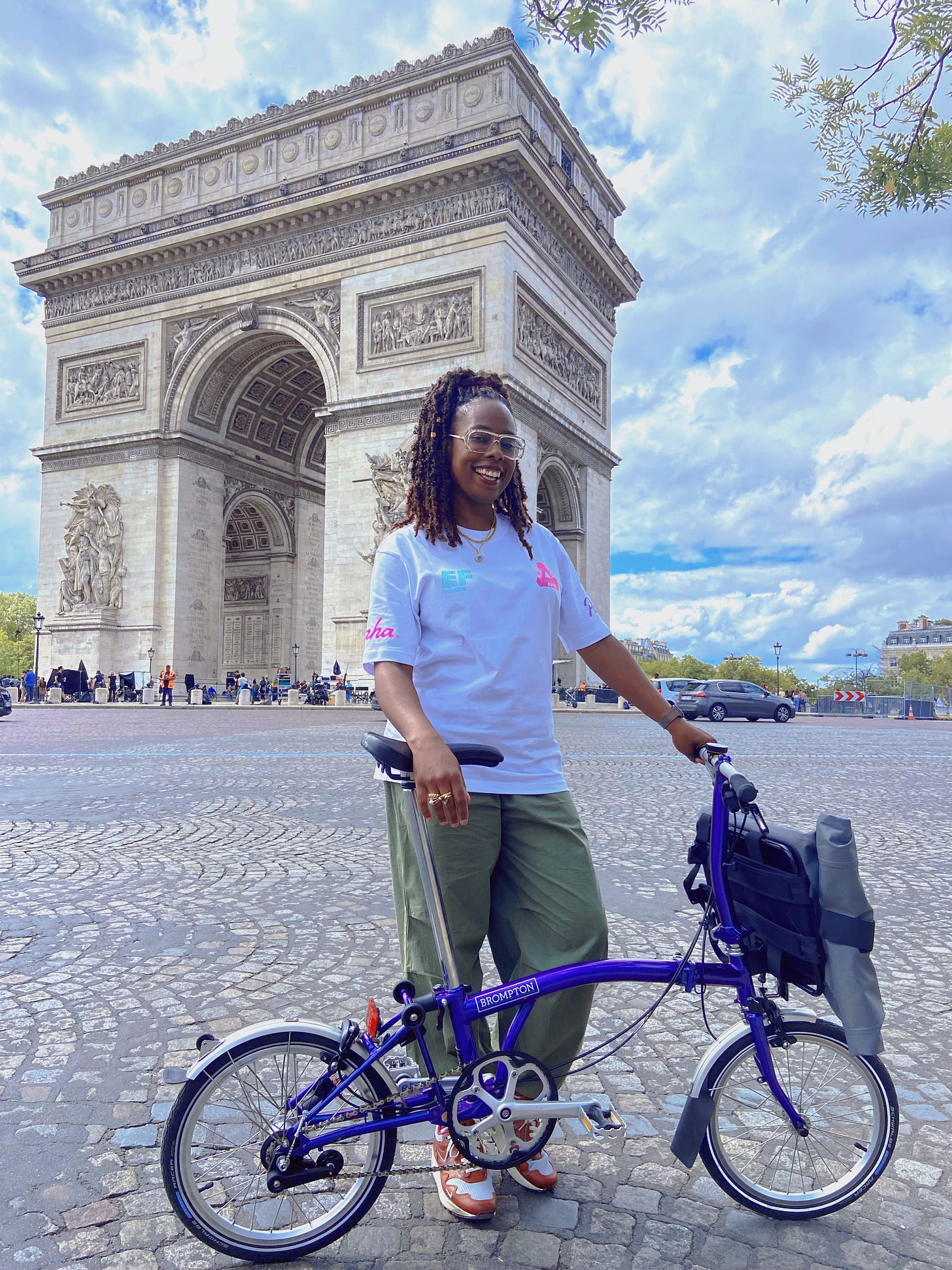 Jools is standing in front of the arc de triomphe in Paris, holding her purple brompton bicycle. She is wearing a white t-shirt and green trousers and smiling. 