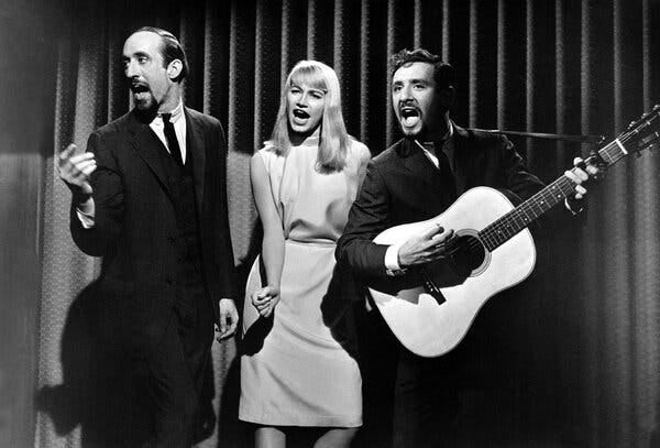 A black-and-white photo of Peter, Paul and Mary in performance. They all have animated expressions on their faces and wide-open mouths. Peter Yarrow, at the right, is playing a guitar; the other two are not playing instruments.