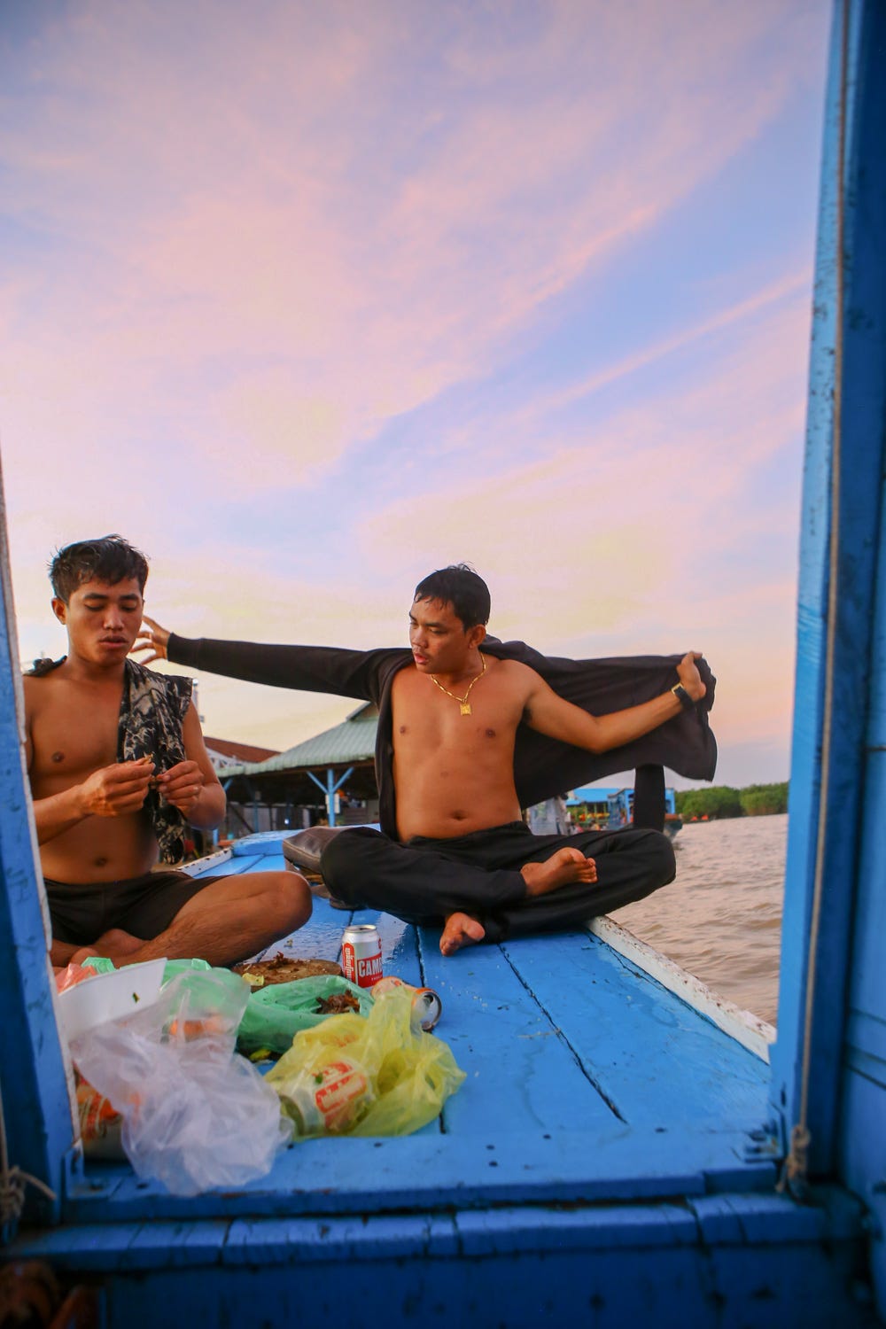 Two young Cambodian men get dressed on the front of a boat with a pink and blue sky in the background. Before them on the deck of the boat sit many plastic bags full of roasted meats.