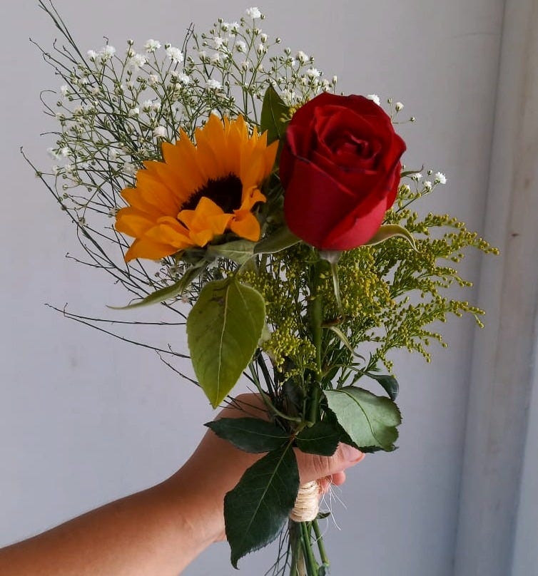 Uma mão segura um buquê de flores simples, com um girassol e uma rosa vermelha