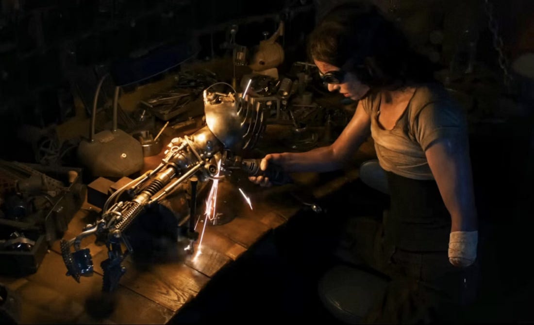 In a movie still, a femme amputee is working on a metal prosthetic arm, sending sparks flying down on a workshop table.