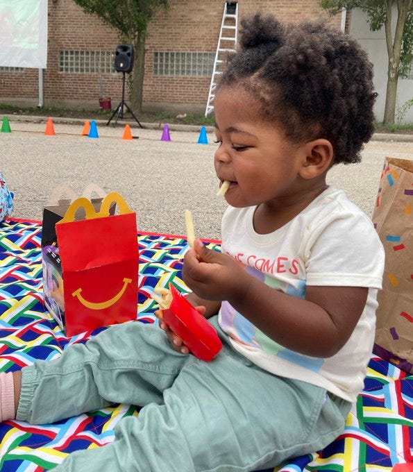 Picture of a Black toddler girl sitting on a multicolored blanket eating a McDonald’s French fries from a happy meal