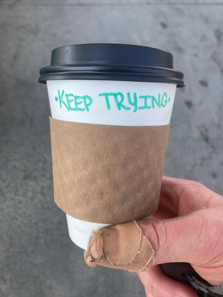 paper coffee cup on which is written in green ink 'Keep Trying.' My thumb is bandaged.