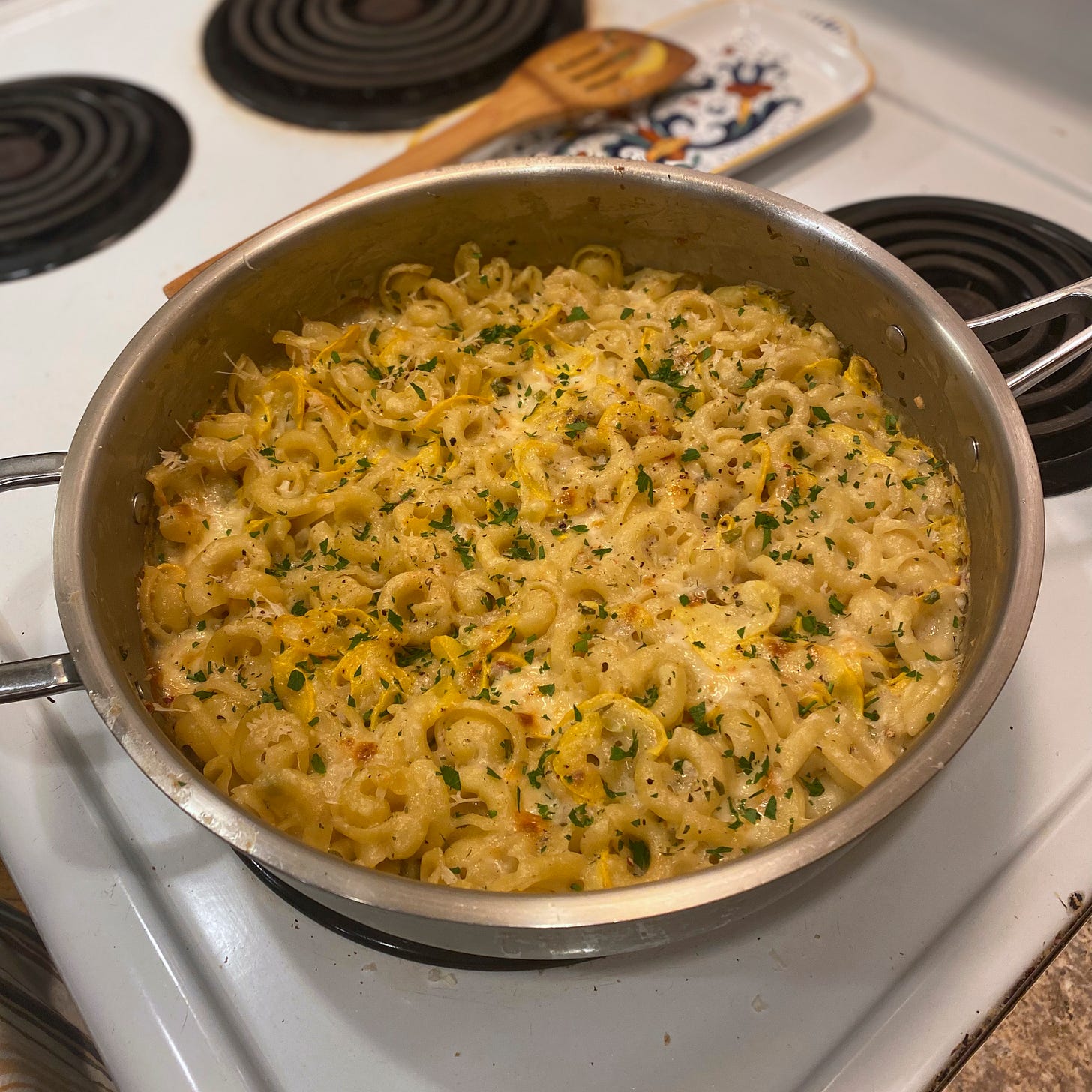 On top of the stove, a large steel pan filled with baked cappelletti in a light-coloured sauce, with pieces of yellow zucchini blending into the dish. Browned bits of cheese are visible at the top and edges, and chopped parsley is scattered across it. A wooden spoon sits on a spoon rest in the background.