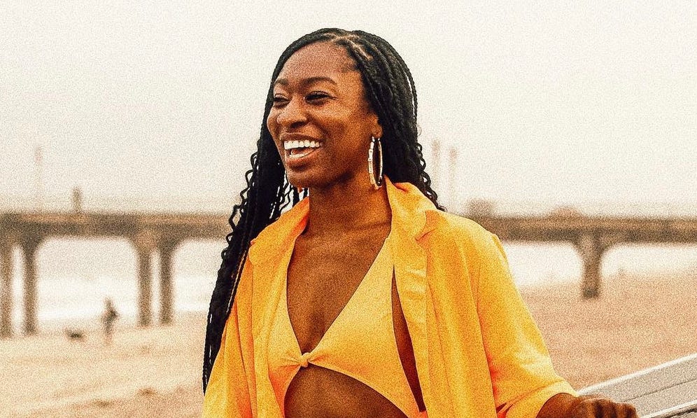 Aaricka is smiling, while wearing a bright yellow swimsuit on the beach.