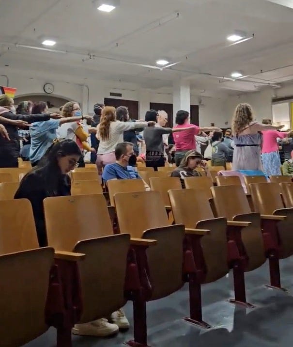 Protesters dancing "Macarena" at CEC board meeting in school auditorium, seen from behind with arms out in dance move.
