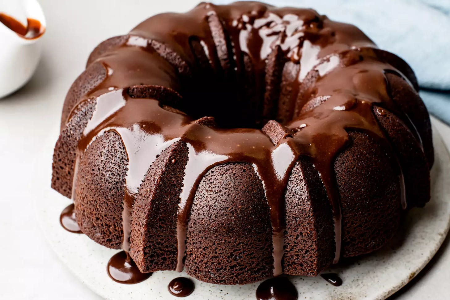 Chocolate glaze being drizzled unevenly over a chocolate Bundt cake, dripping down the sides onto plate