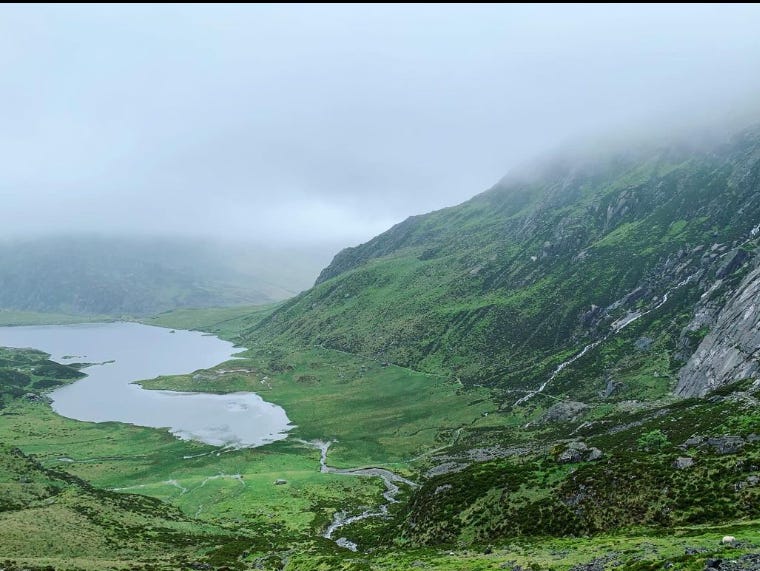 Looking out from a hillside across a mountain valley with an oblong lake and low-hanging misty clouds in the distance