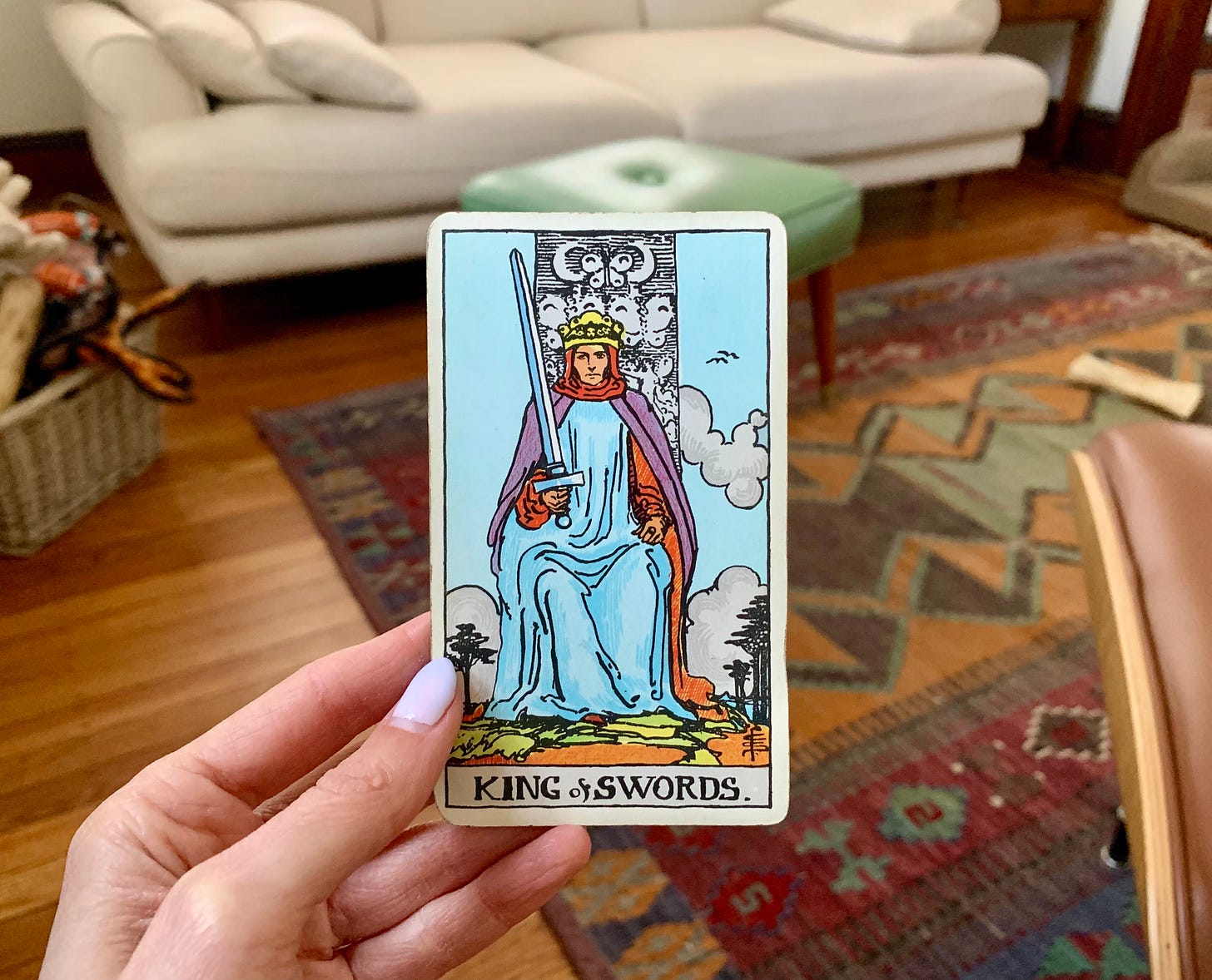 A hand is holding a tarot card, King of Swords by Pamela Colman Smith. In the image a person is dressed in blue, purple, and red and sitting on a throne. He's holding a sword which is standing straight up toward the sky. 