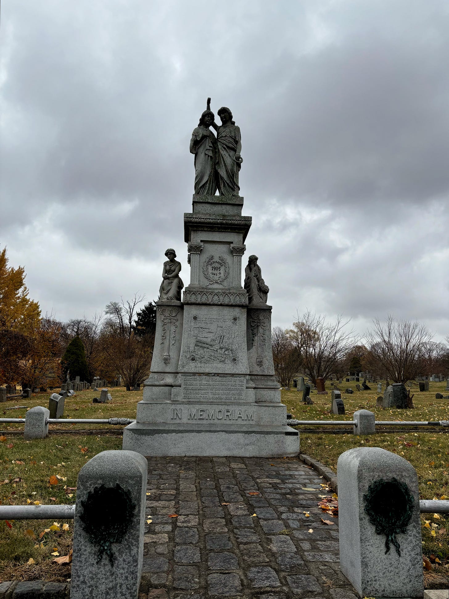 The full memorial at All Faiths. Over the engraving two angelic figures hold one another. Beneath the engraving in large letters it says IN MEMORIAM. In smaller letters it reads: Erected by the organization of the General Slocum Survivors and the Public. In memory of the 6 unidentified dead who lost their lives on the steamboat General Slocum June 15, 1904.