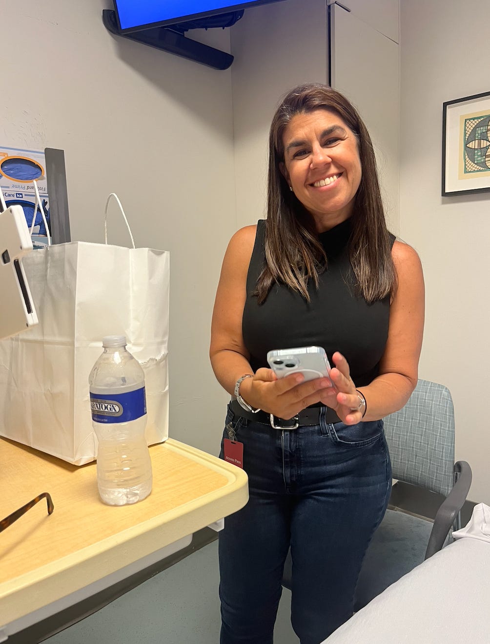 Dr. Dara Kass stands in Amber's hospital room. She looks into the camera smiling broadly. She holds her cell phone. On the table next to her is a white take-out food bag.