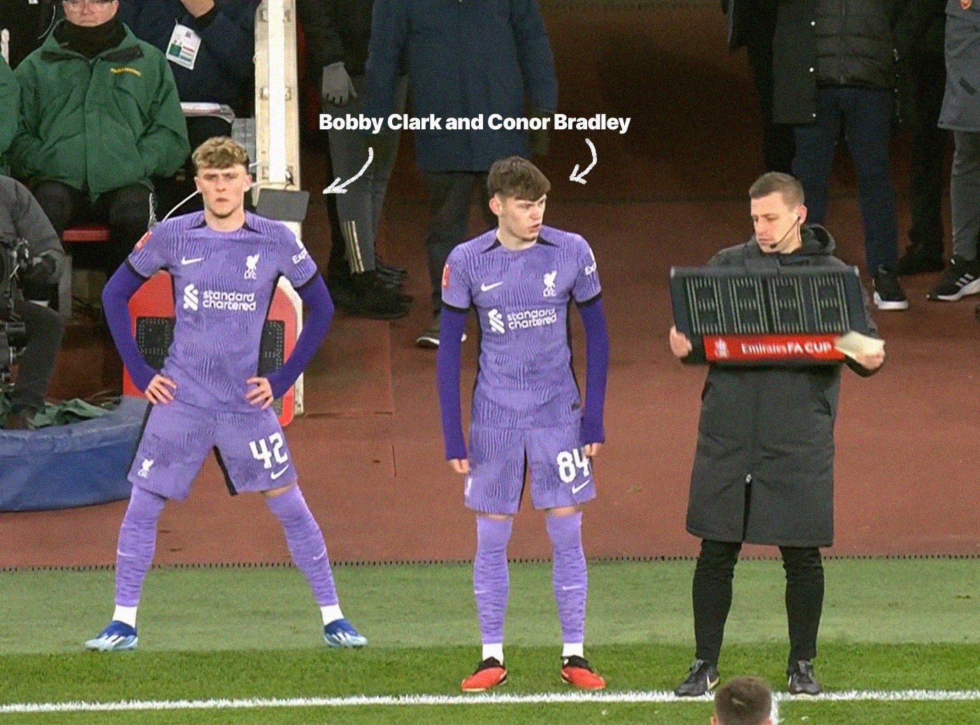 A screenshot of Bobby Clark and Conor Bradley on the sideline, preparing to come on as susbtitutes in an FA Cup fixture against Arsenal. They're both wearing purple Liverpool kits. The fourth official, wearing a black coat and holding the substitution board, is stood beside them.