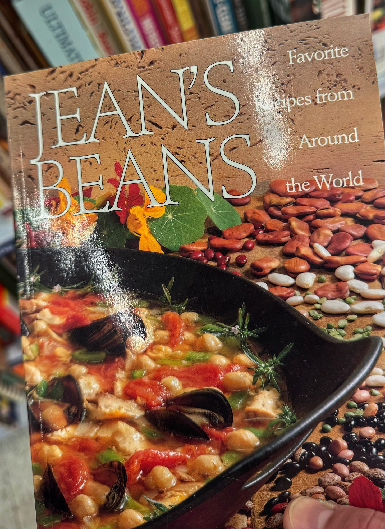 The cover of a cookbook called "Jean's Beans: Favorite Recipes from Around the World" held up in front of a shelf of books in a thrift store