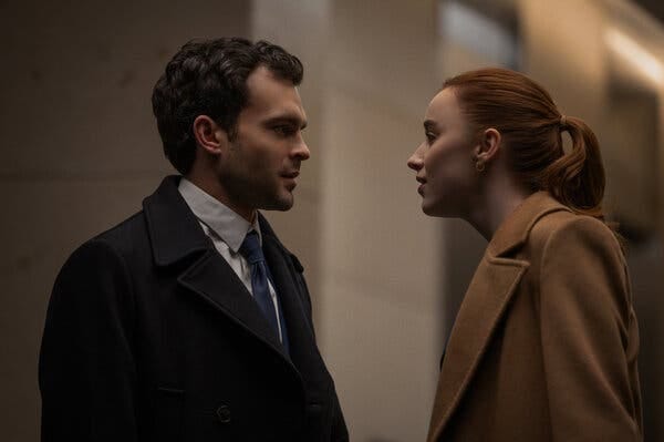 A man and a woman, him in a dark coat and her in a tan coat, stand and have a conversation. 