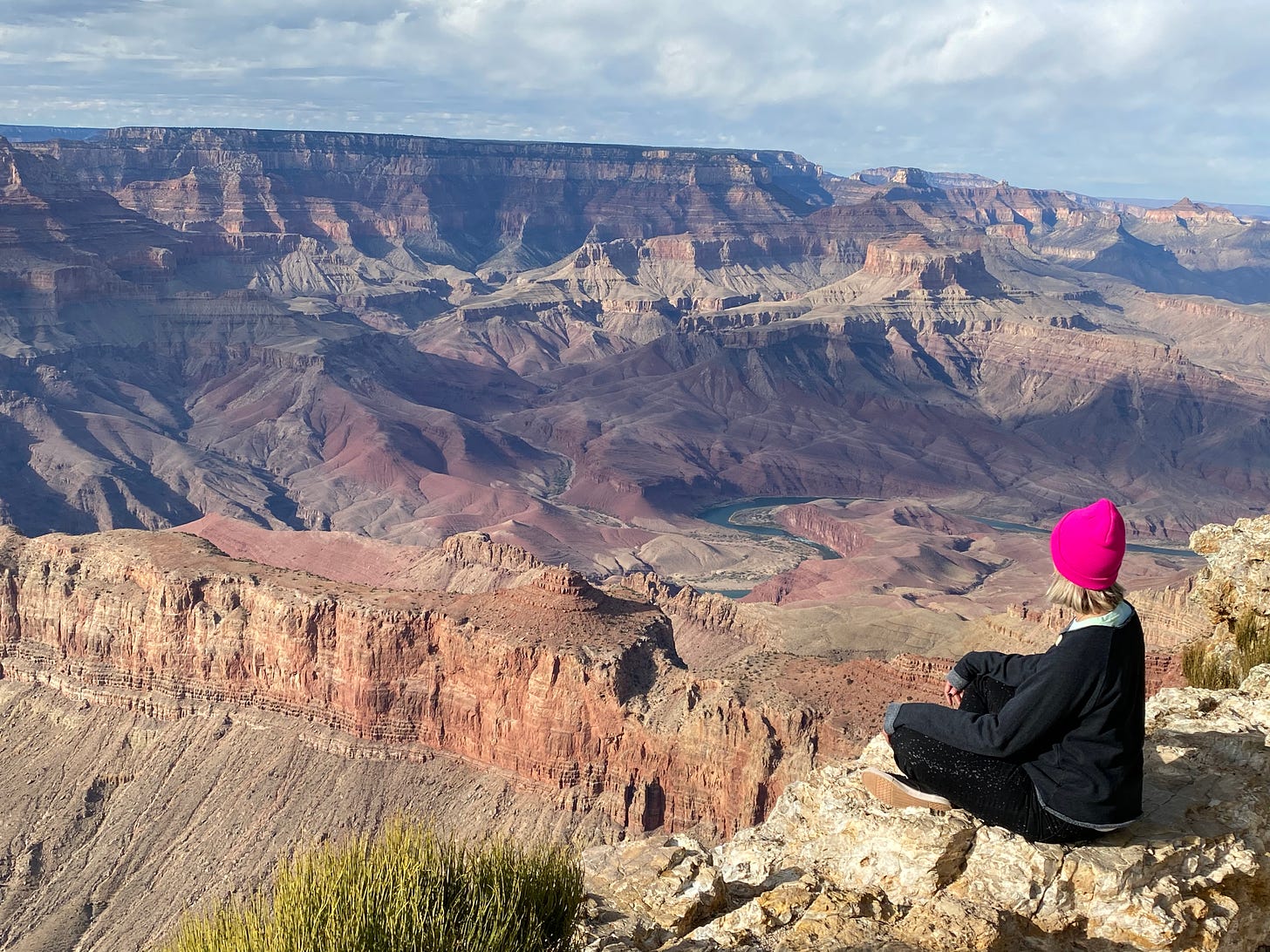 A woman in a pink beanie sits on the rim of the Grand Canyon, looking out on its expanse below.