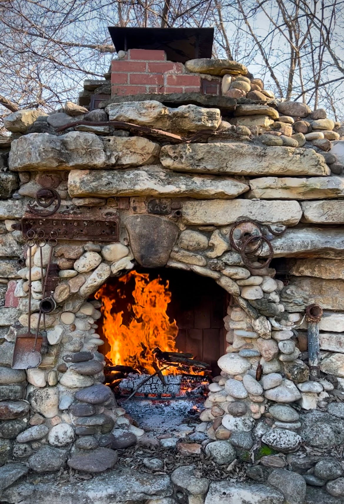 Color photo of outdoor fireplace made from concrete debris, metal trash, and river rock, with a blazing fire on