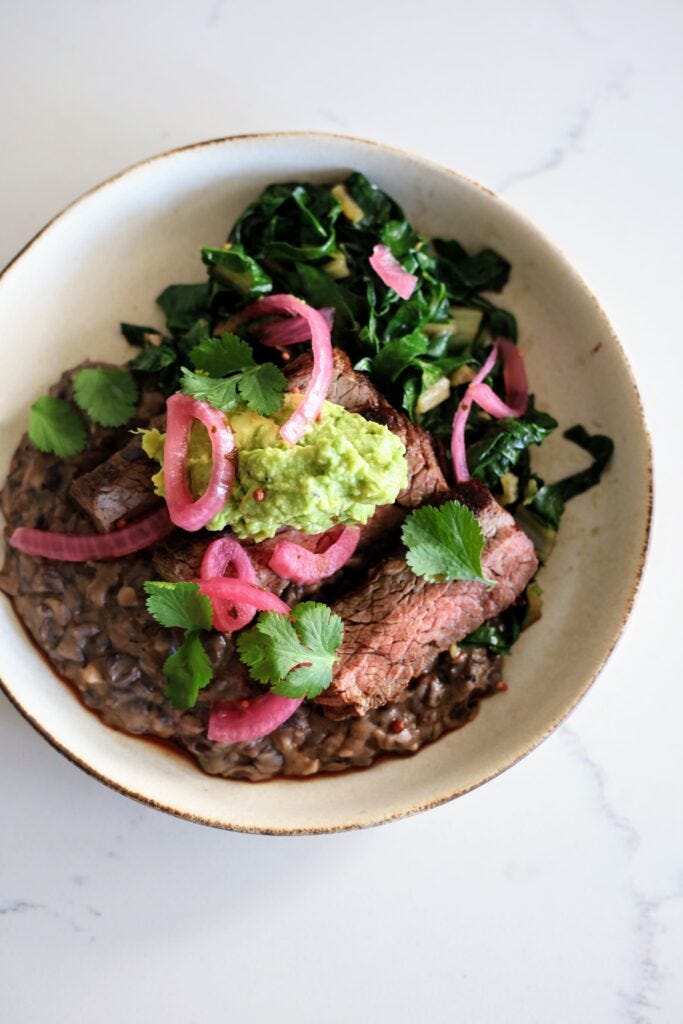 close up of a completed bowl of mashed beans on the bottom, with steak, topped with smased avocado, pickled red onions, and cilantro with a side of cooked chard 