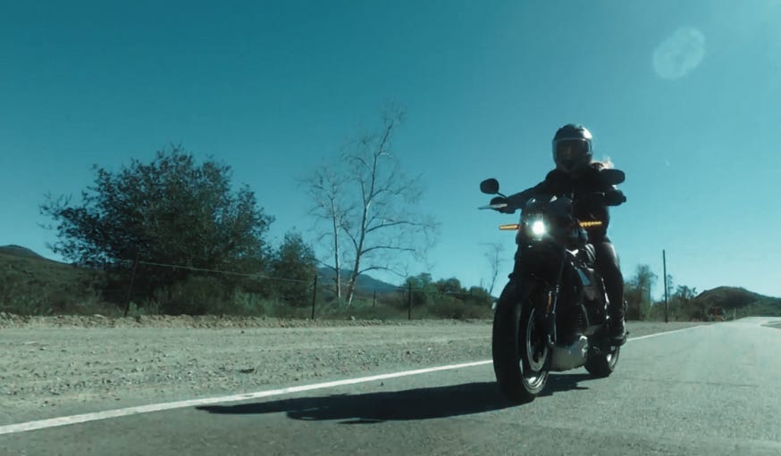 The woman races down the road on her motorcycle. The sky is clear and the road stretches out behind her. Her face is obscured.