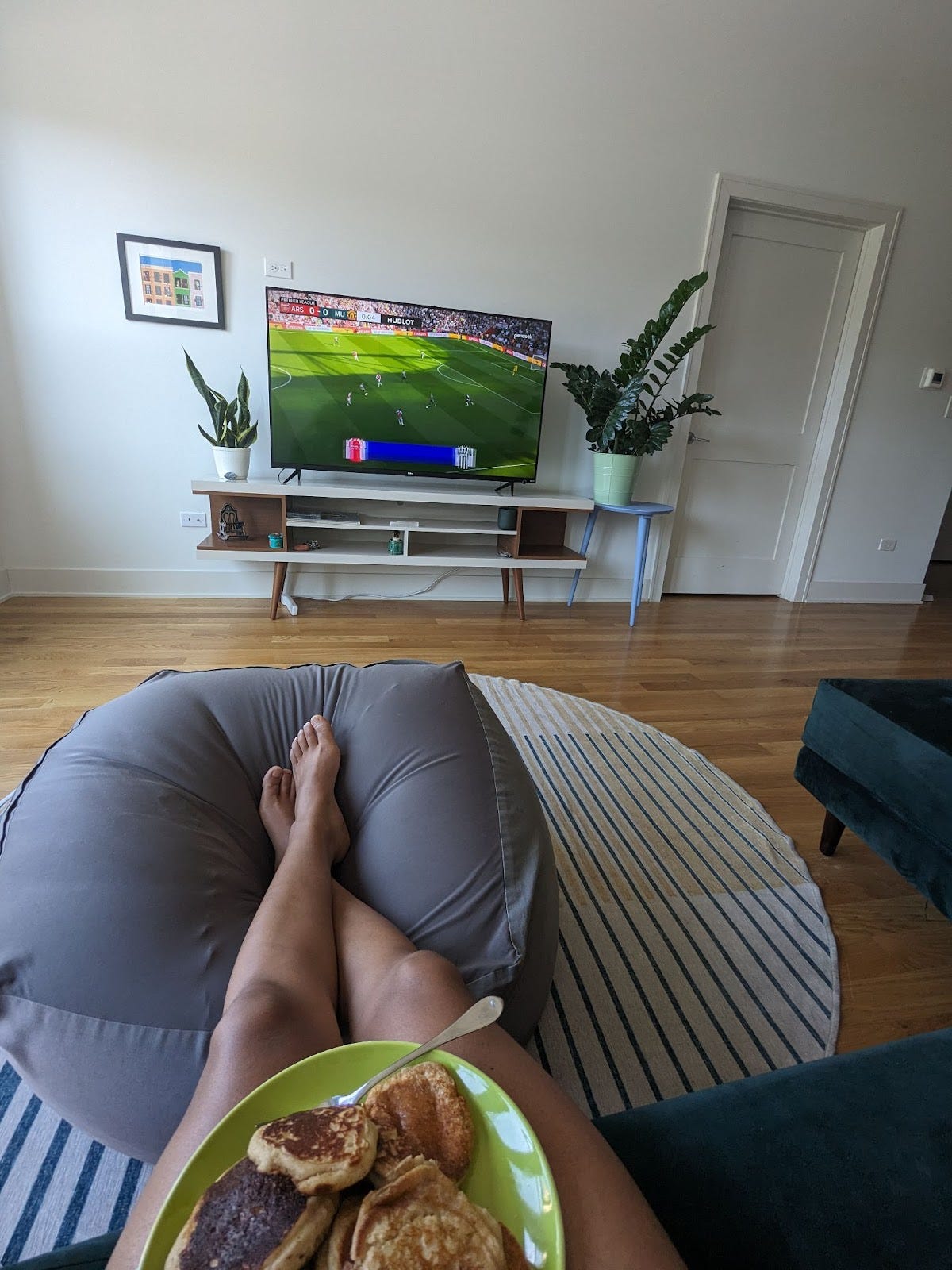 A soccer game is displayed on flat screen TV. Nathalie props her feet on a gray beanbag chair and has a plate of pancakes on her thighs to enjoy while she watches the game