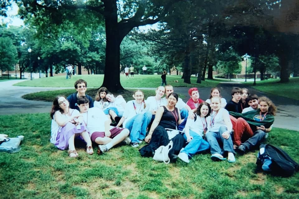 A photograph of about 20 teens sitting in a big pile on a bit of grass with a college campus in the background.