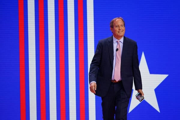 Ken Paxton stands before a screen that has red and white vertical stripes and a large white star on a blue background.