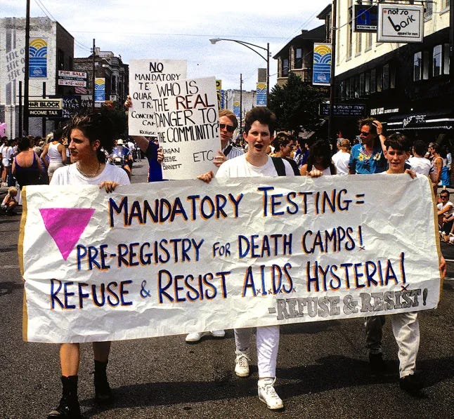 A group of activists march down a city street during a protest, holding a large banner that reads: “Mandatory Testing = Pre-Registry for Death Camps! Refuse & Resist A.I.D.S. Hysteria!” The banner features a large pink triangle and the logo for Refuse & Resist. Behind them, another sign reads, “No mandatory testing or quarantine. Who is the real danger to the community?” The street is lined with people and buildings A group of activists march down a city street during a protest, holding a large banner that reads: “Mandatory Testing = Pre-Registry for Death Camps! Refuse & Resist A.I.D.S. Hysteria!” The banner features a large pink triangle and the logo for Refuse & Resist. Behind them, another sign reads, “No mandatory testing or quarantine. Who is the real danger to the community?” The street is lined with people and buildings