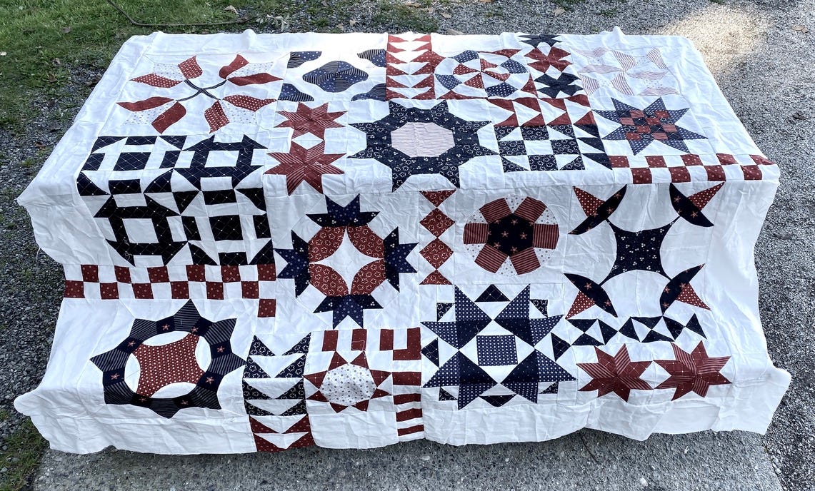 A 65"by 78" quilt top laid on top of a picnic table. There are 21 blocks with different patterns using a variety of fabrics in blues and reds and whites, all on the theme of "stars and stripes".