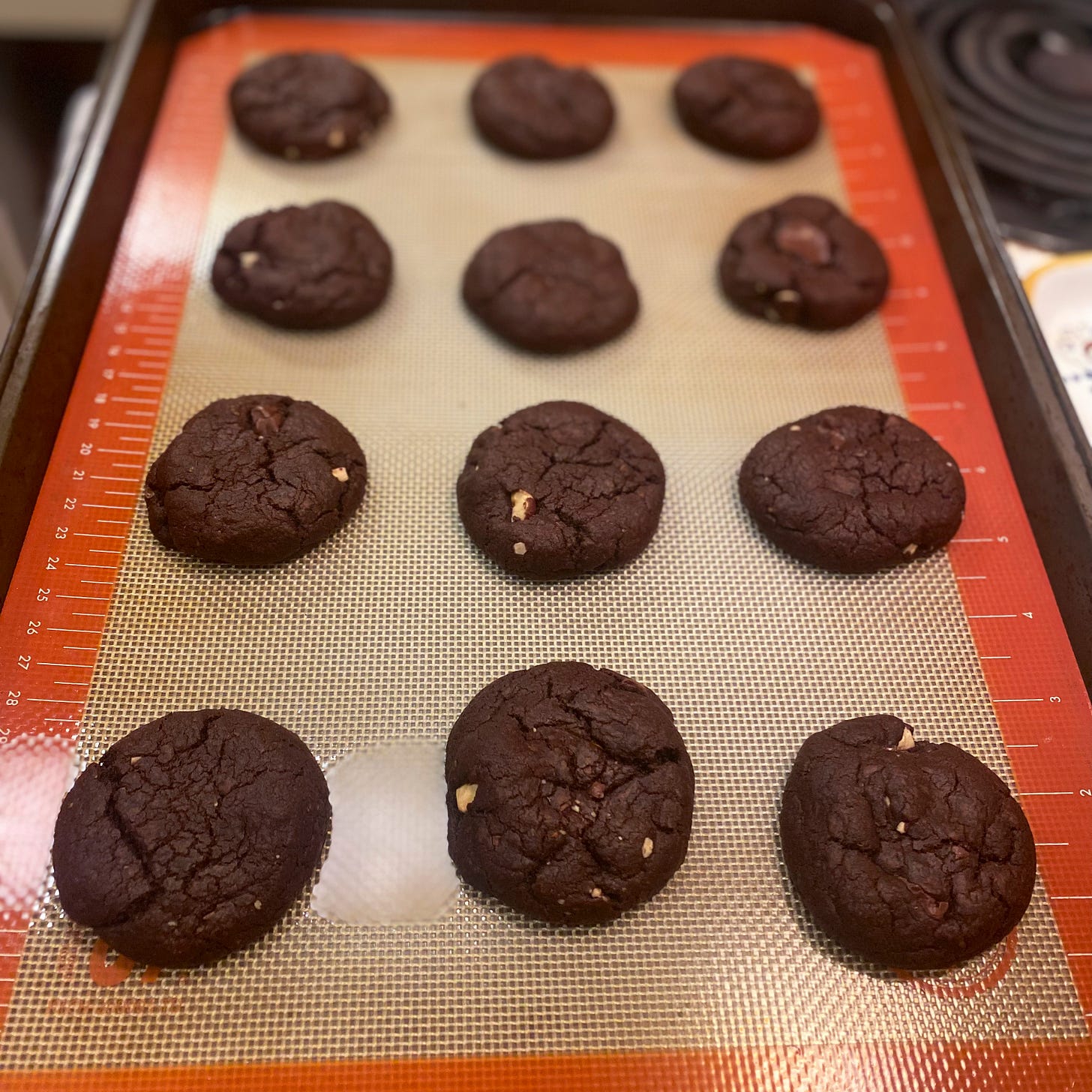 On a baking sheet lined with a silpat, a dozen double chocolate cookies with bits of hazelnut visible throughout. 