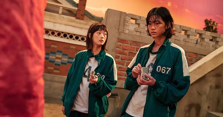 Two people in green tracksuits with numbers 210 and 067 holding cups, stand in a brick courtyard at sunset, looking pensive.