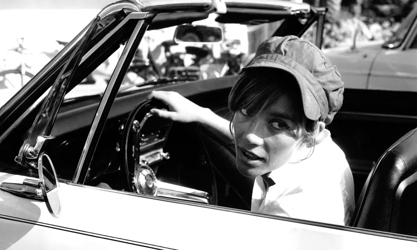 Françoise Hardy dirigindo um carro conversível ainda jovem. Ela usa um boné, tem cabelos presos e está parada olhando para o lado 